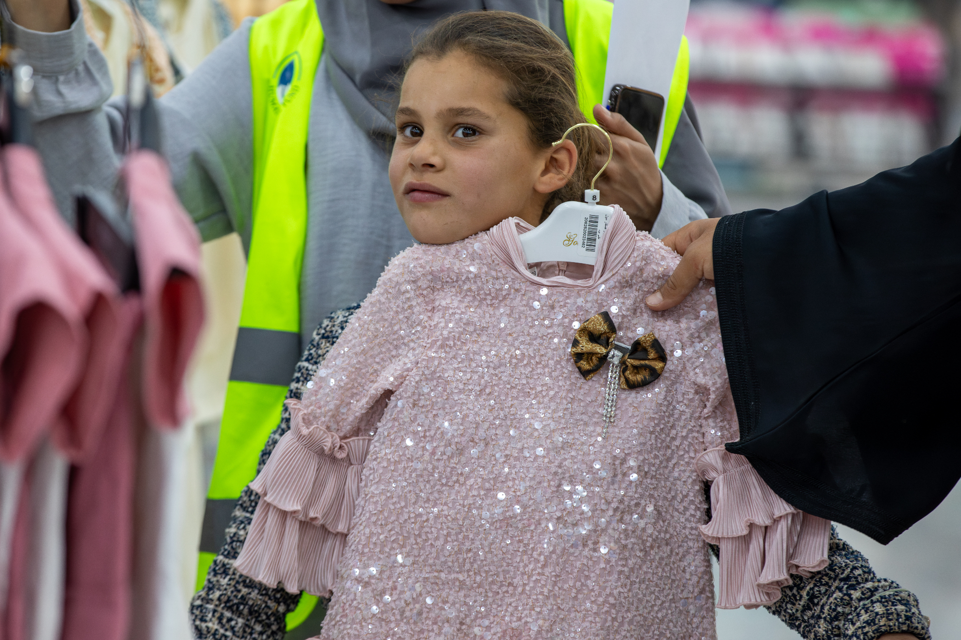 A young girl holding up a pink sequined Eid dress alongside an HDF volunteer in Al Marfaq, Jordan.