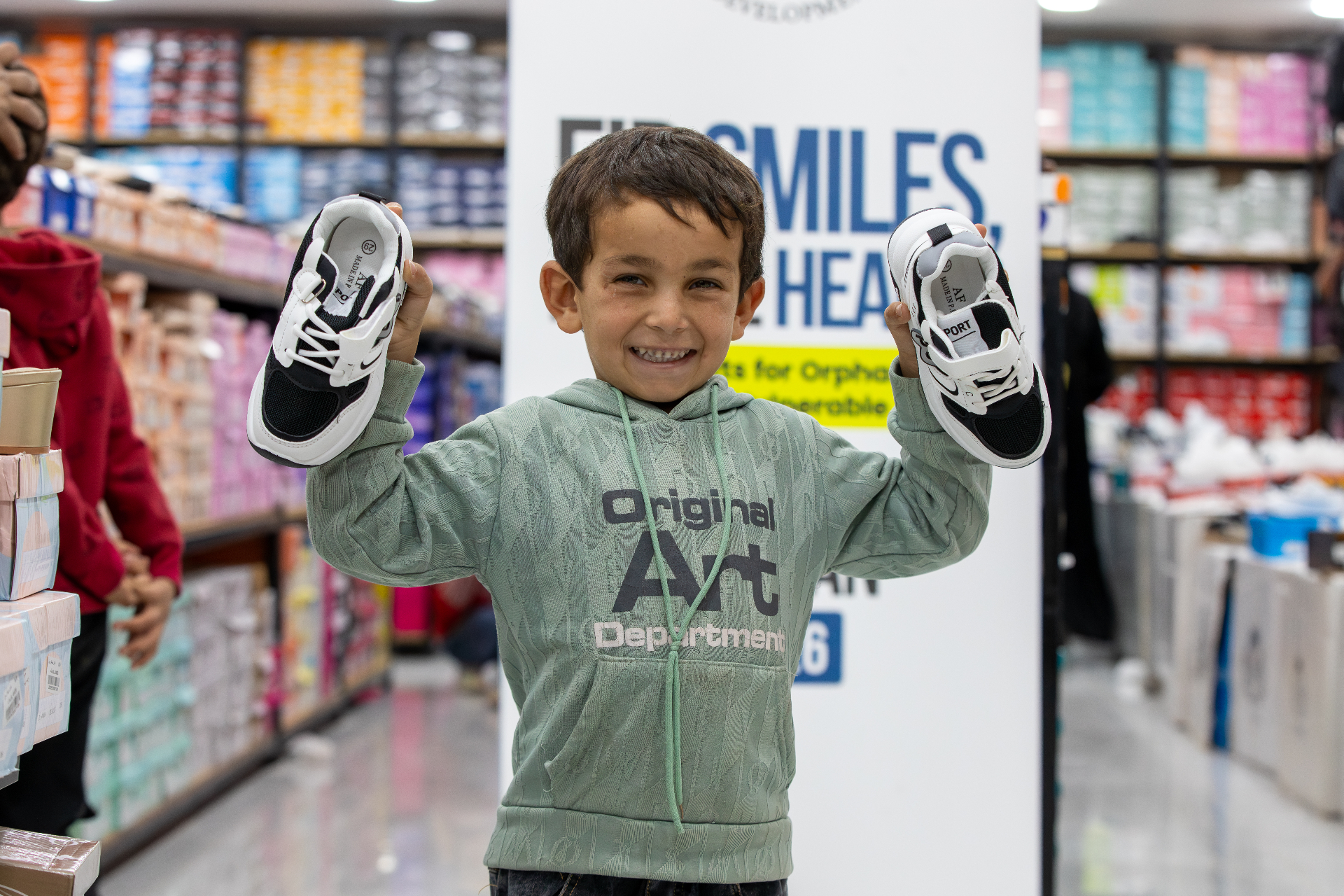 A child smiling and holding new shoes received during HDF's pre-Eid shopping program in Al Marfaq, Jordan.