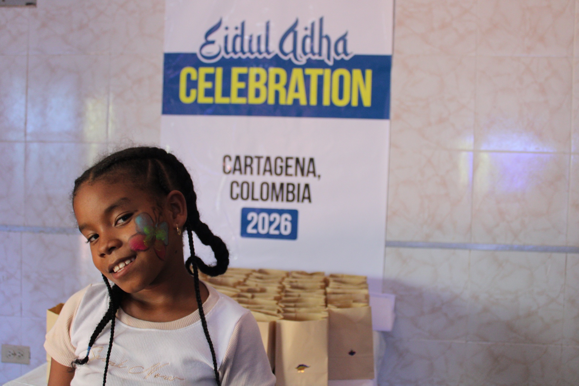 A young girl with face paint smiling in front of an HDF Eidul Adha Celebration 2026 banner in Cartagena, Colombia.