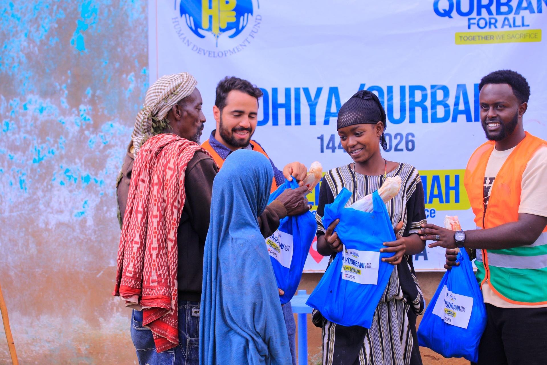 HDF field workers in orange vests handing blue Udhiya / Qurbani meat share bags to a young woman and an elderly man beside a 'Qurbani For All — Feed the Ummah, Ethiopia' banner.