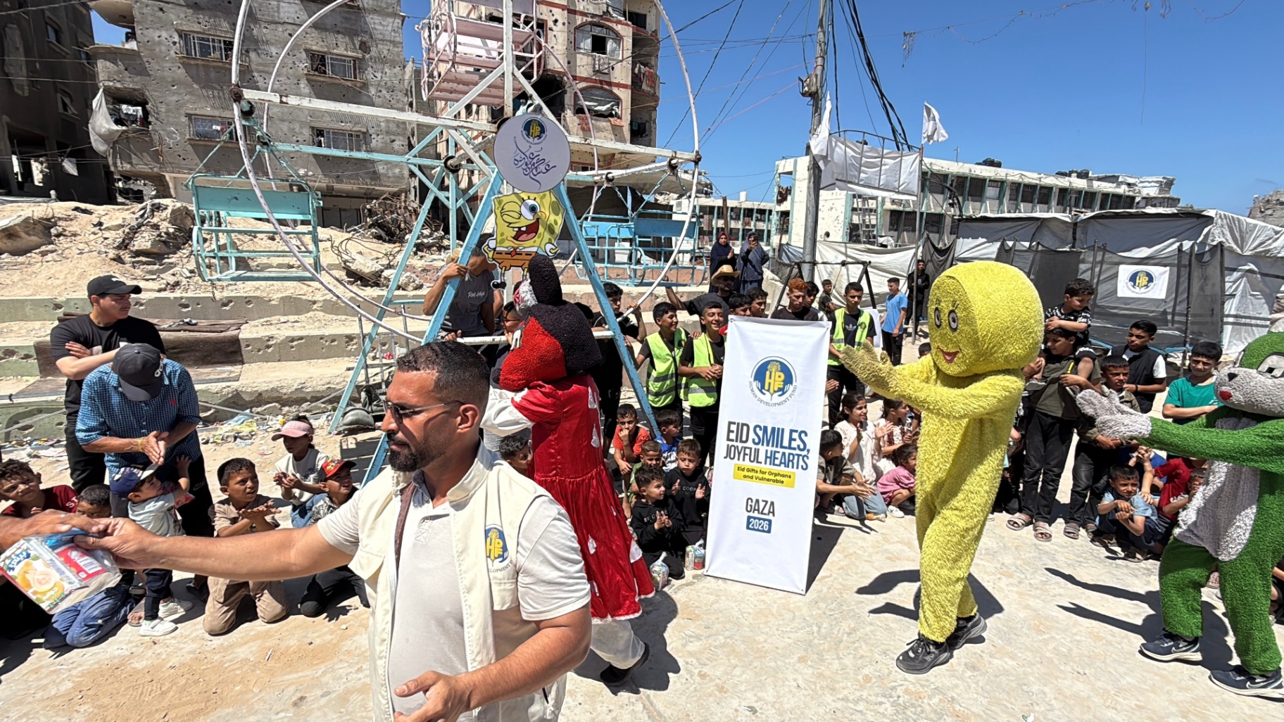 Costumed mascots performing in front of a small Ferris wheel at HDF's Eid Smiles, Joyful Hearts celebration in Gaza.