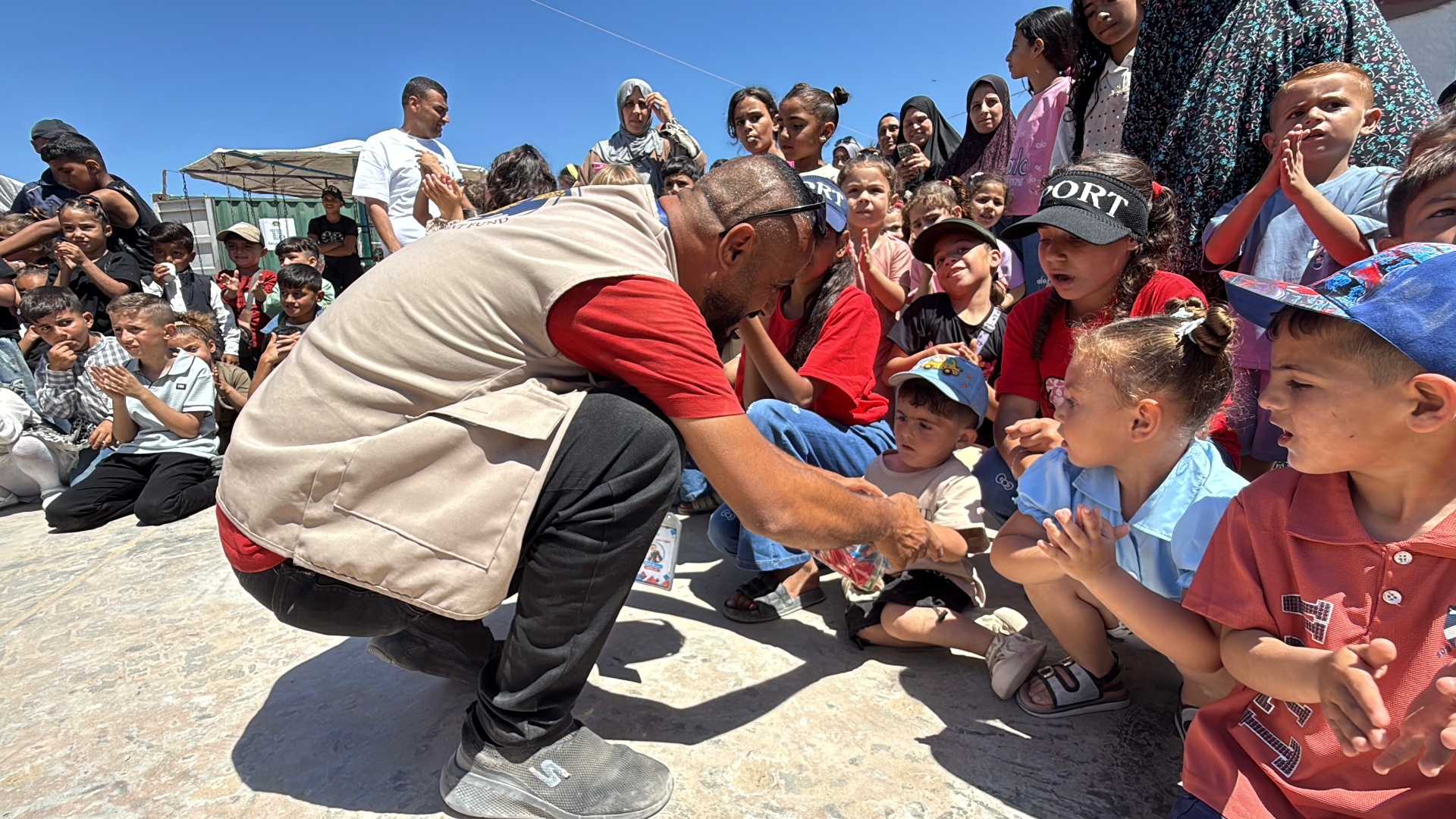 An HDF team member kneeling down to hand Eid gifts to young children clapping and gathered around him in Gaza.
