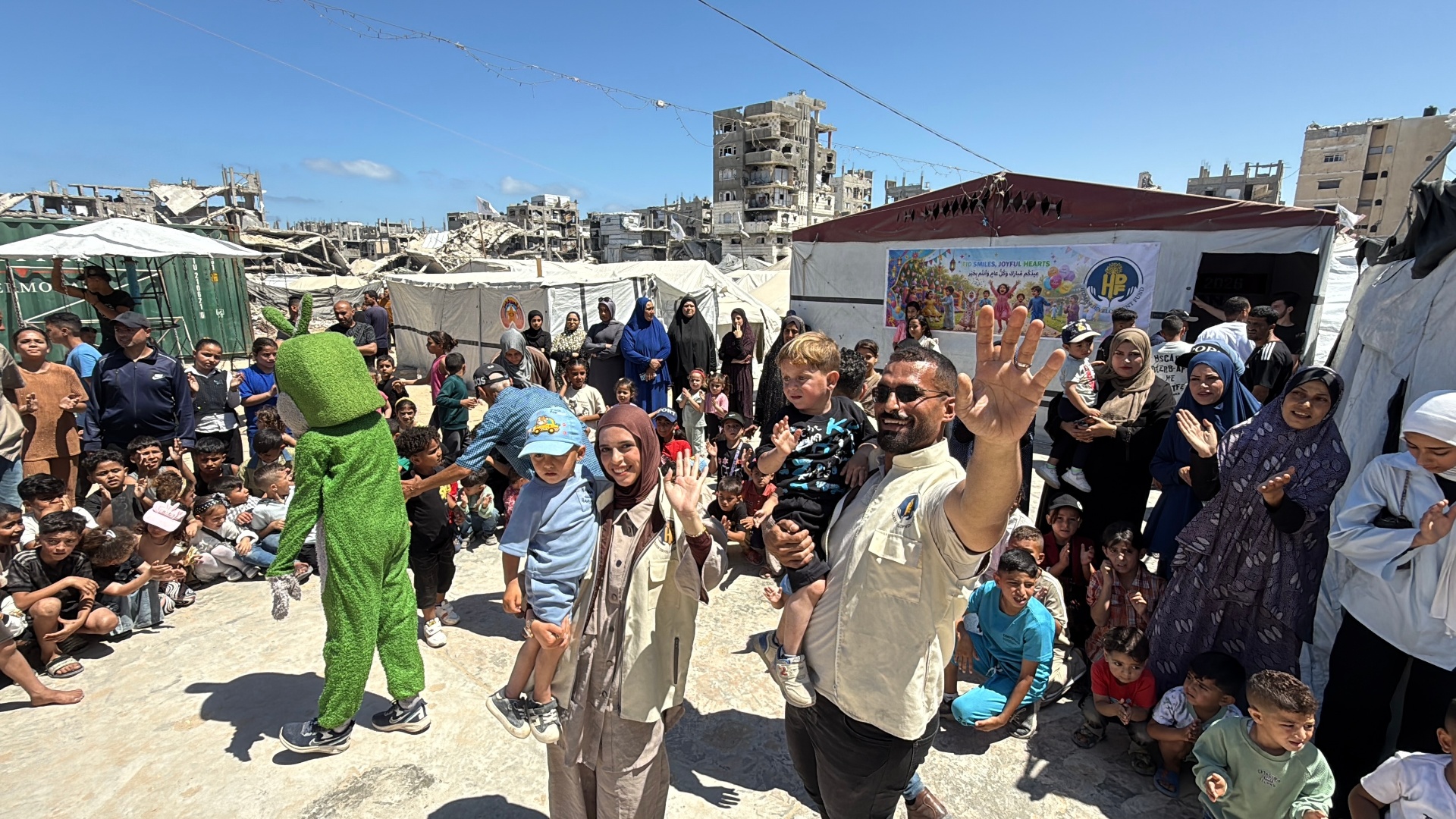 HDF team members waving with children in front of the Eid Smiles, Joyful Hearts banner in Gaza.