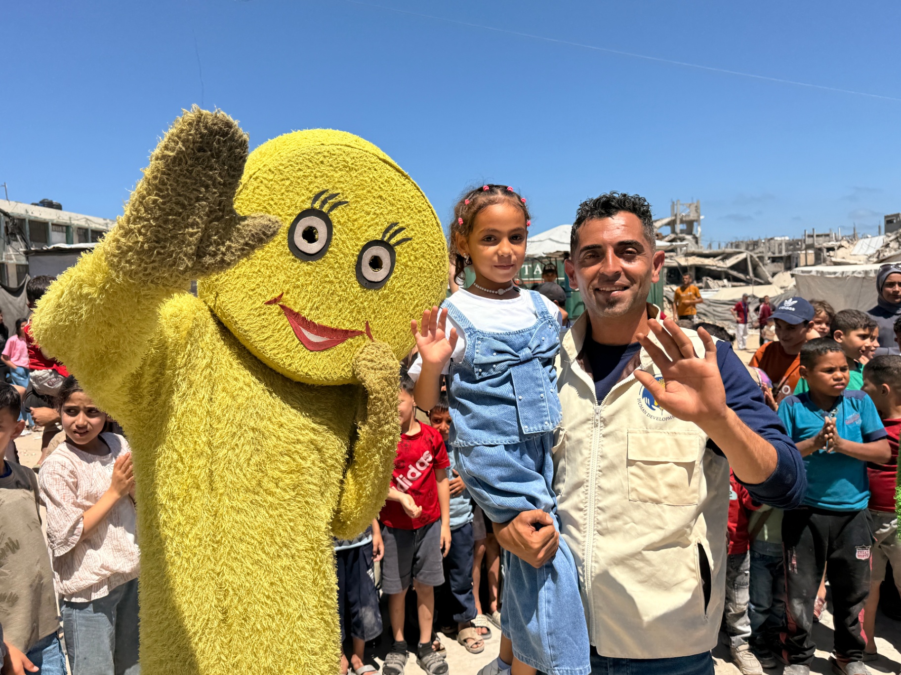 A girl waving alongside a yellow mascot and an HDF team member at the Eid celebration in Gaza.