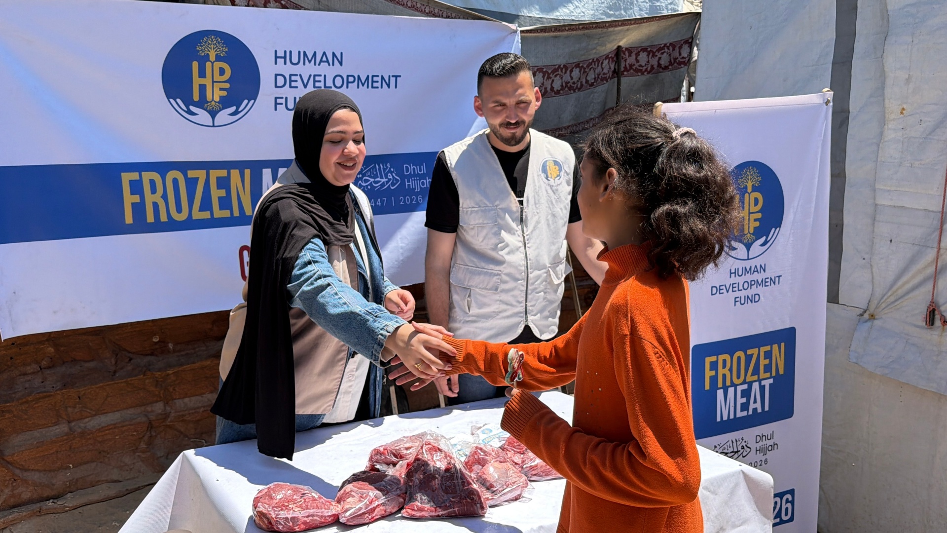 A young girl in an orange sweater reaching out to receive her family's Frozen Meat Qurbani share from a smiling HDF field worker in a hijab beside an HDF Frozen Meat — Gaza banner.