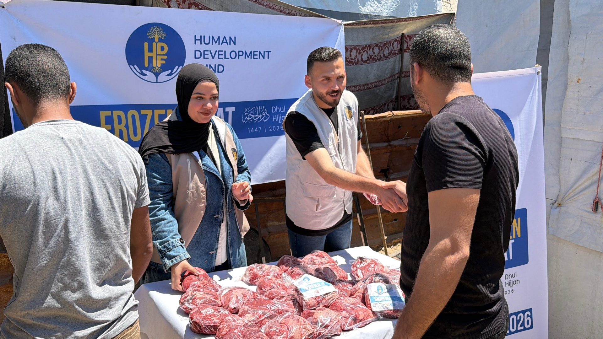 An HDF field worker in a branded vest shaking hands with a man receiving Frozen Meat shares as another beneficiary waits beside a table of vacuum-sealed Qurbani packs in Gaza.