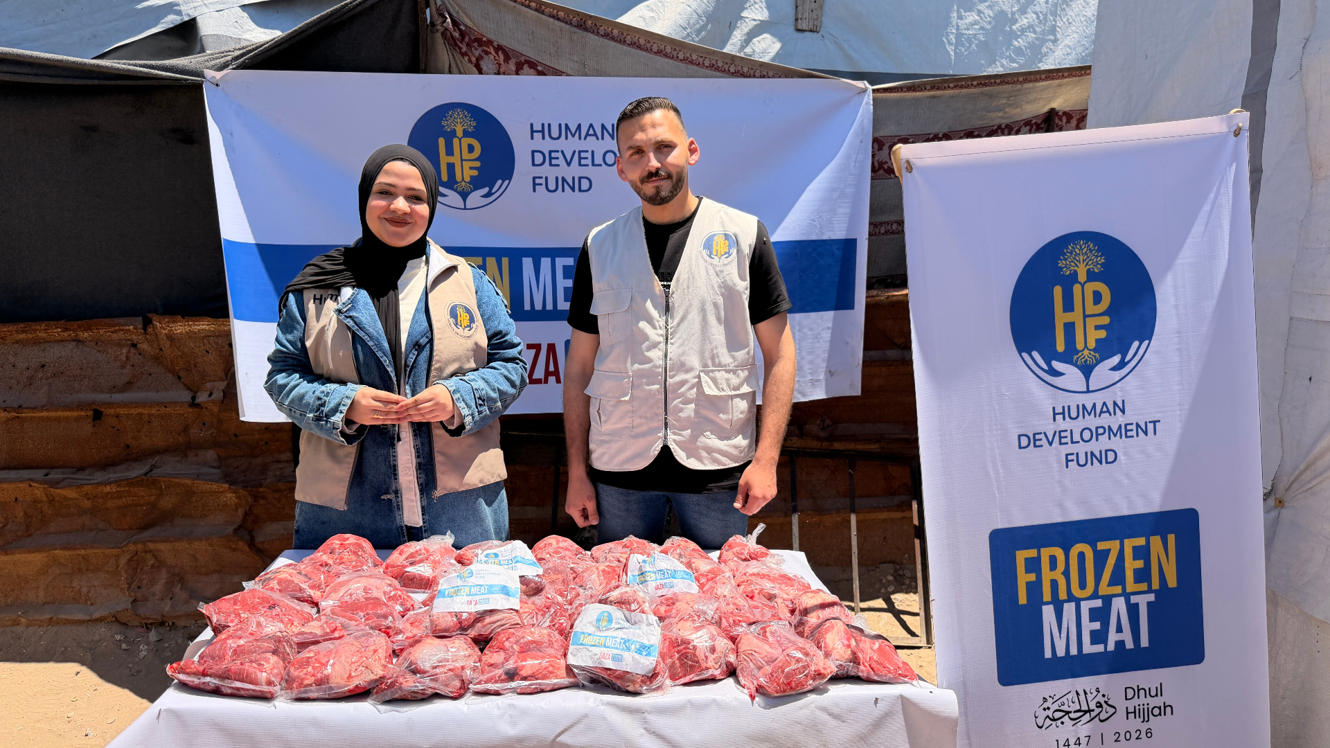 Two HDF field workers in branded vests standing behind a table stacked with vacuum-sealed Frozen Meat Qurbani shares in front of an HDF Frozen Meat — Gaza banner.