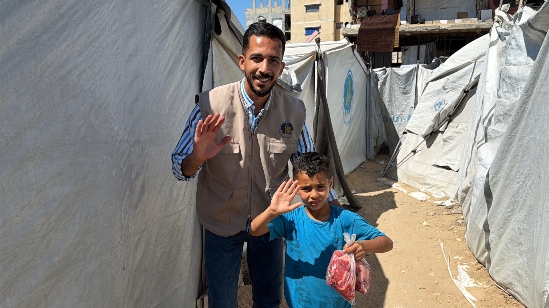 A young boy in a blue shirt smiling and holding up a bag of Frozen Meat next to an HDF field worker in a tan vest in a Gaza displacement camp.