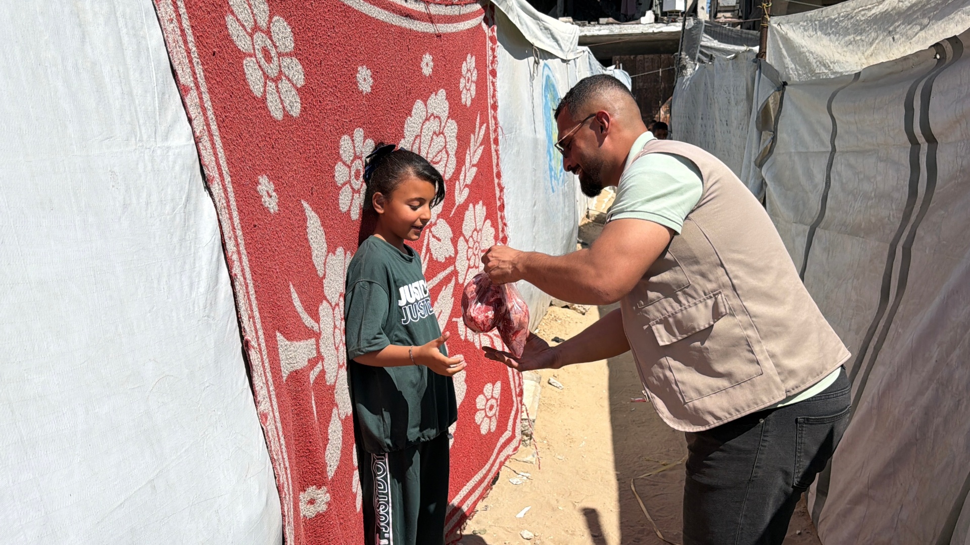 An HDF field worker in a tan vest handing a wrapped bag of Frozen Meat to a young girl standing beside a red and white patterned blanket between displacement tents in Gaza.