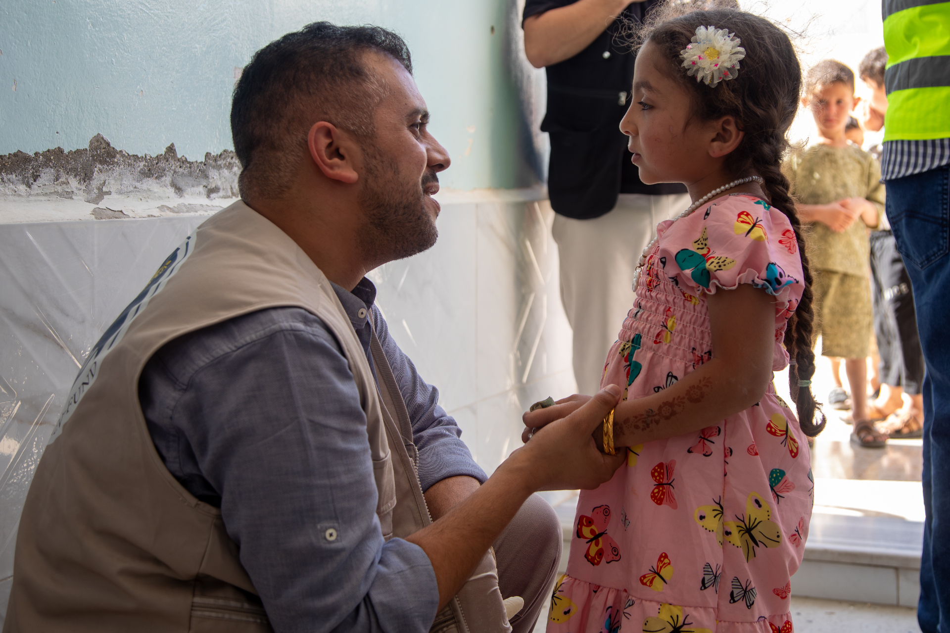 An HDF field worker kneeling to greet a young girl in a pink butterfly dress during the Qurbani distribution in Jordan.