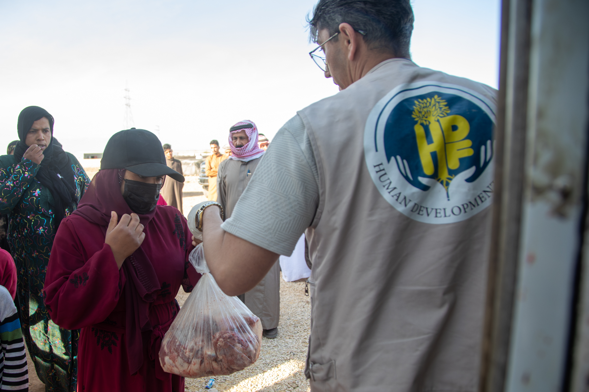 An HDF field worker in a branded vest handing a wrapped Qurbani meat share to a woman in a red abaya as community members wait behind her.