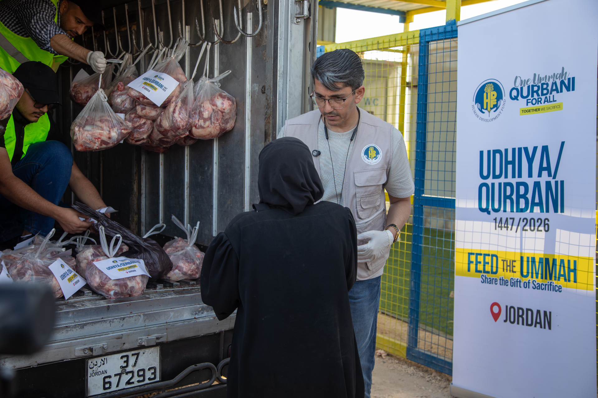 An HDF field worker in gloves preparing labeled Qurbani meat shares from a distribution truck beside a 'Feed the Ummah — Jordan' banner.