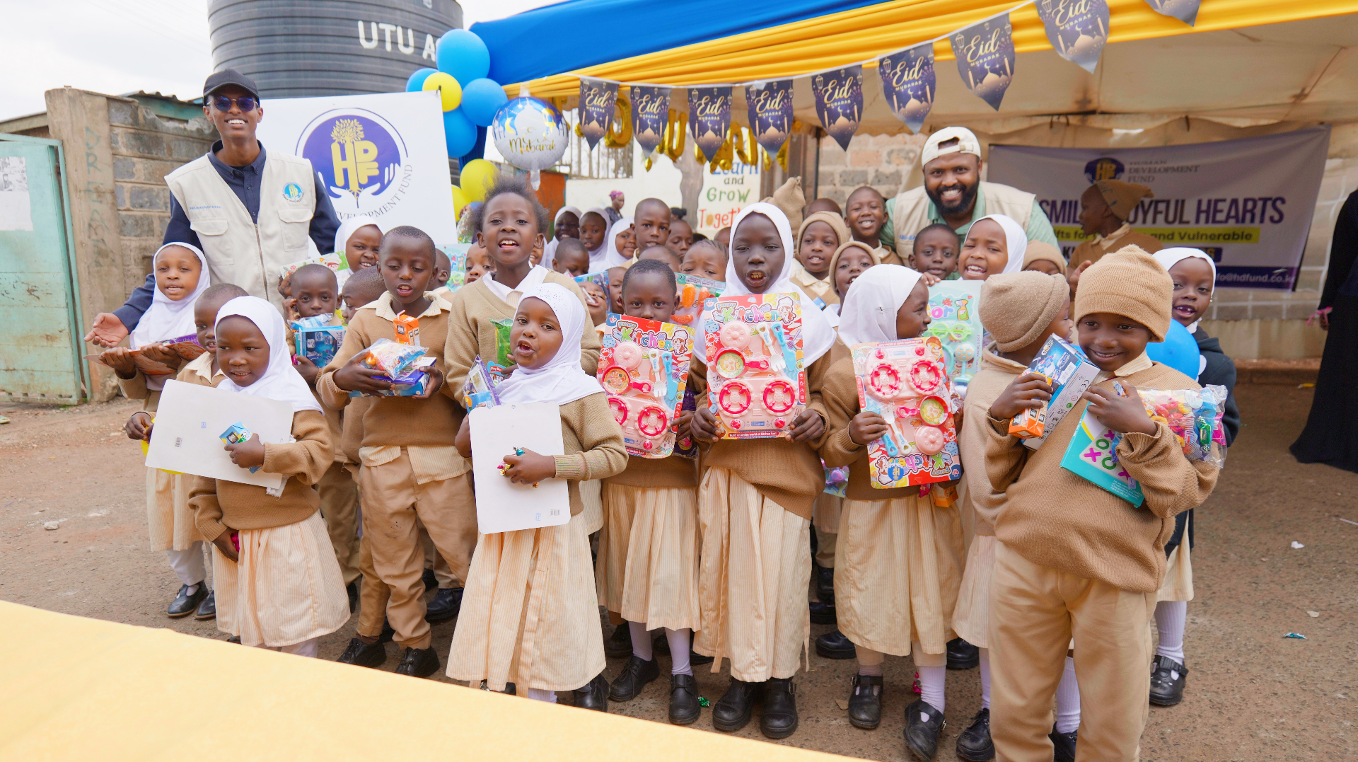 A large group of smiling children holding Eid gifts alongside HDF team members under a decorated Eid Mubarak tent in Kenya.