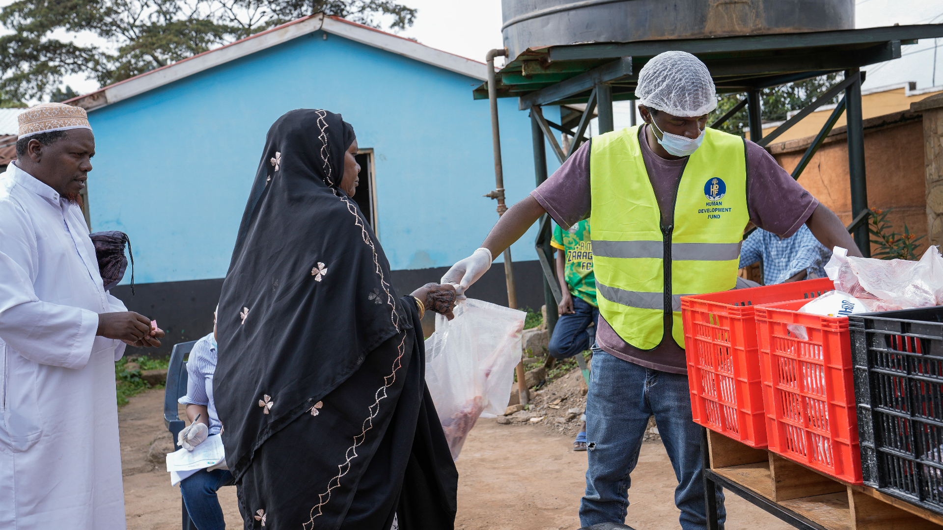 An HDF volunteer in a yellow vest and gloves handing a wrapped Qurbani meat share from a red crate to a woman in a black embroidered hijab as a local imam looks on in Kenya.