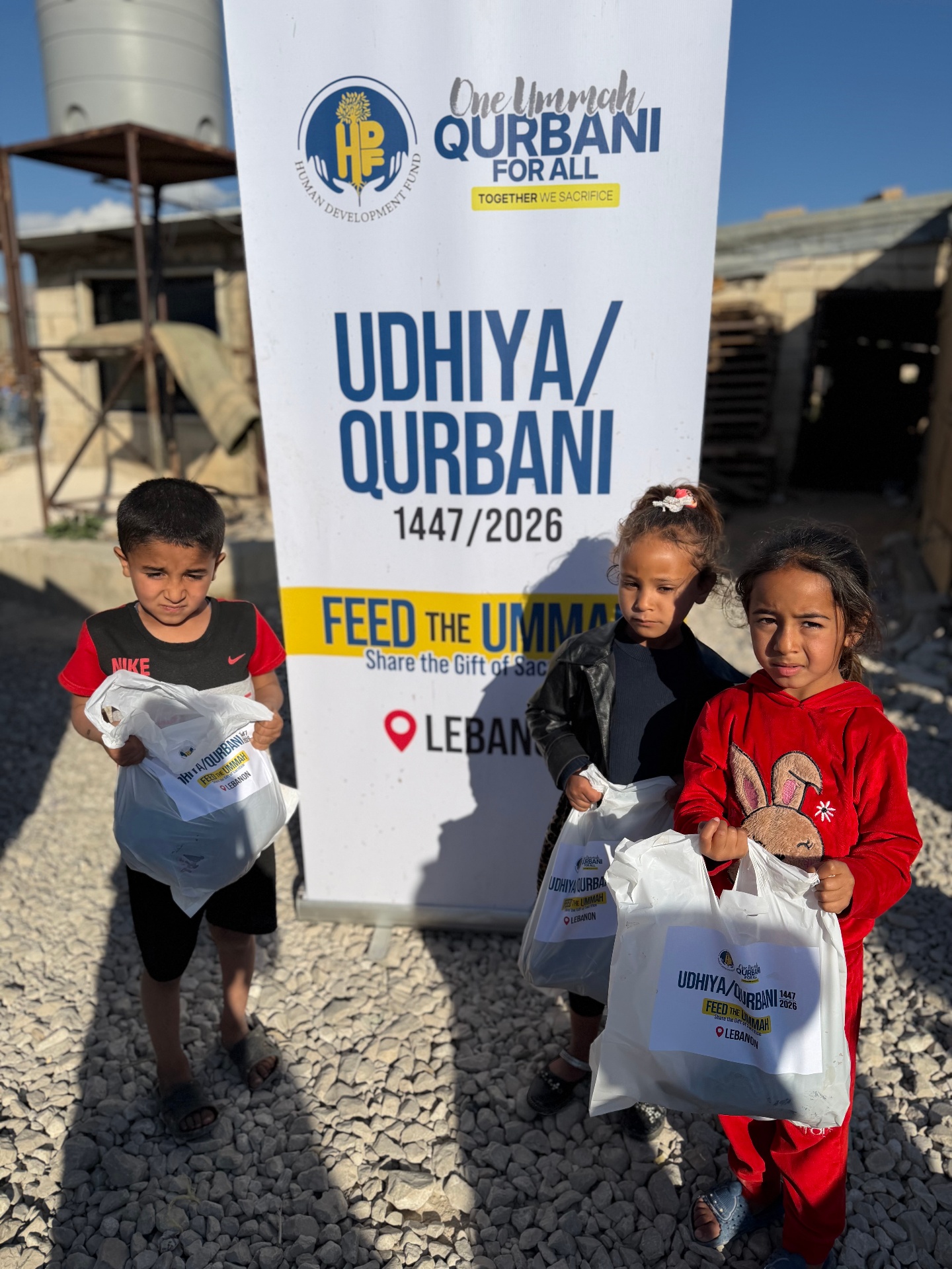 Three young children holding wrapped Udhiya/Qurbani meat share bags beside a One Ummah Qurbani For All — Lebanon banner during HDF's 1447/2026 distribution.