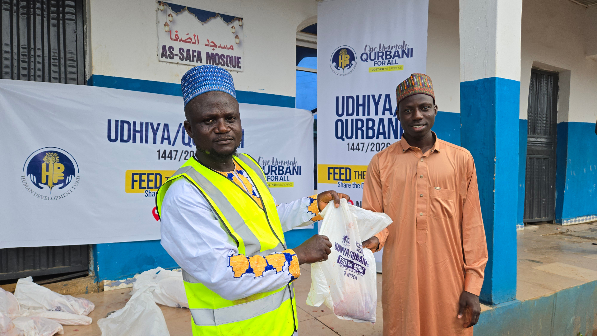 An HDF volunteer in a high-vis vest handing a Qurbani meat bag to a young man in an orange thobe in front of the As-Safa Mosque and Udhiya / Qurbani — Feed the Ummah banners in Nigeria.