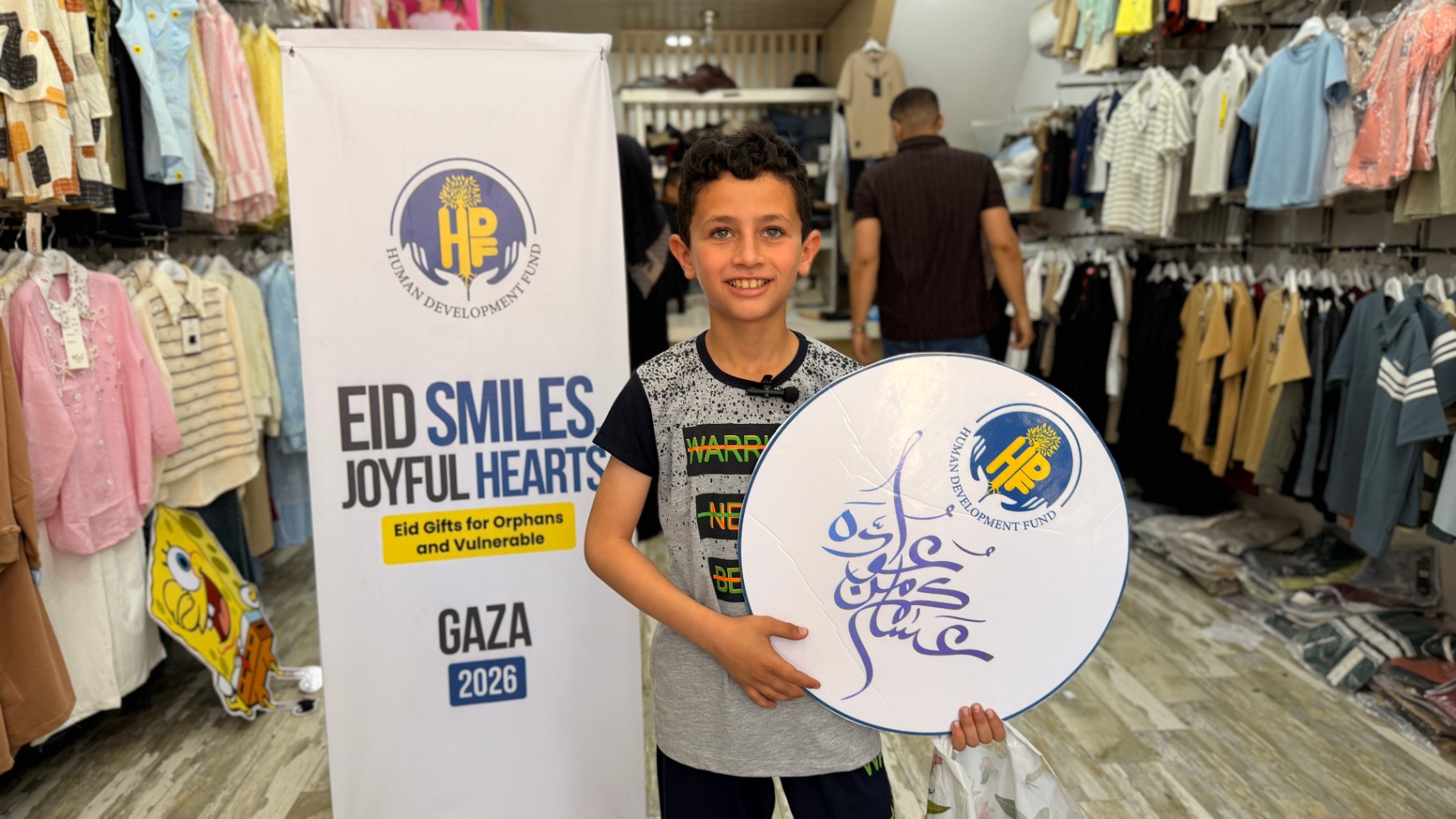 A boy holding an HDF Eid sign and his shopping bag inside a clothing store in Gaza.