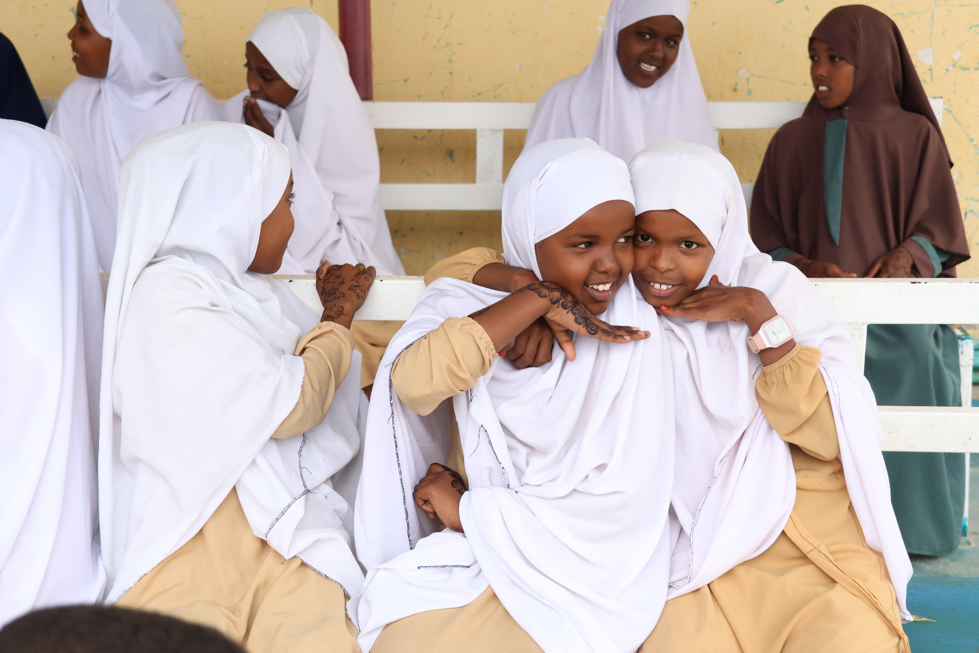 Two young schoolgirls in white hijabs and tan uniforms embracing with bright smiles and henna-decorated hands at HDF's Eid celebration in Somalia.