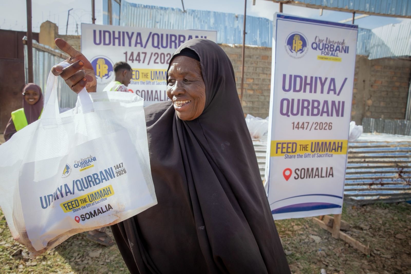 An elderly Somali woman in a dark hijab smiling and lifting her white 'Udhiya / Qurbani — Feed the Ummah, Somalia' meat share bag in front of HDF's One Ummah Qurbani For All banners.