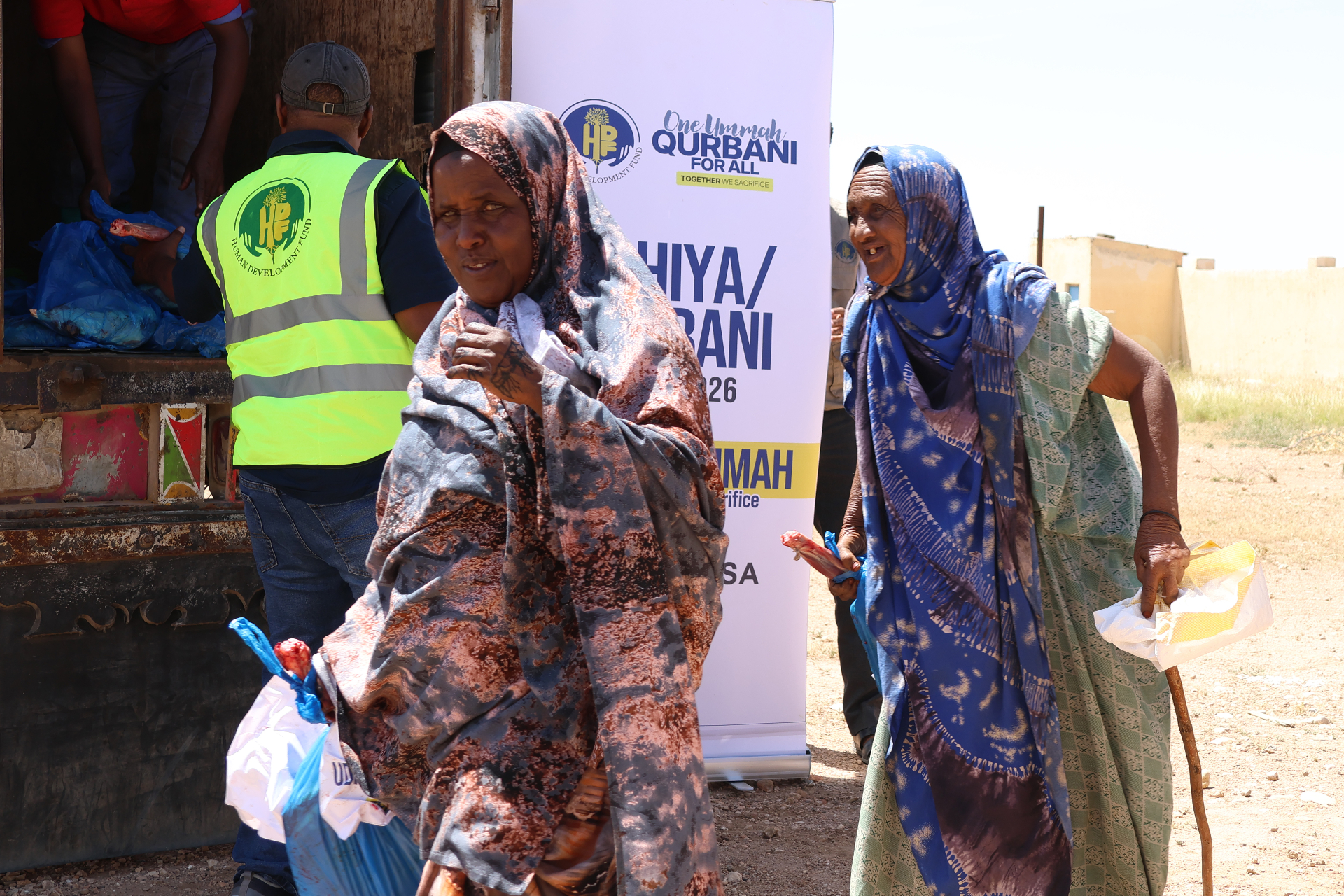 Two elder Somali women in colorful shawls carrying their One Ummah Qurbani For All meat share bags away from the HDF distribution truck in Somalia.