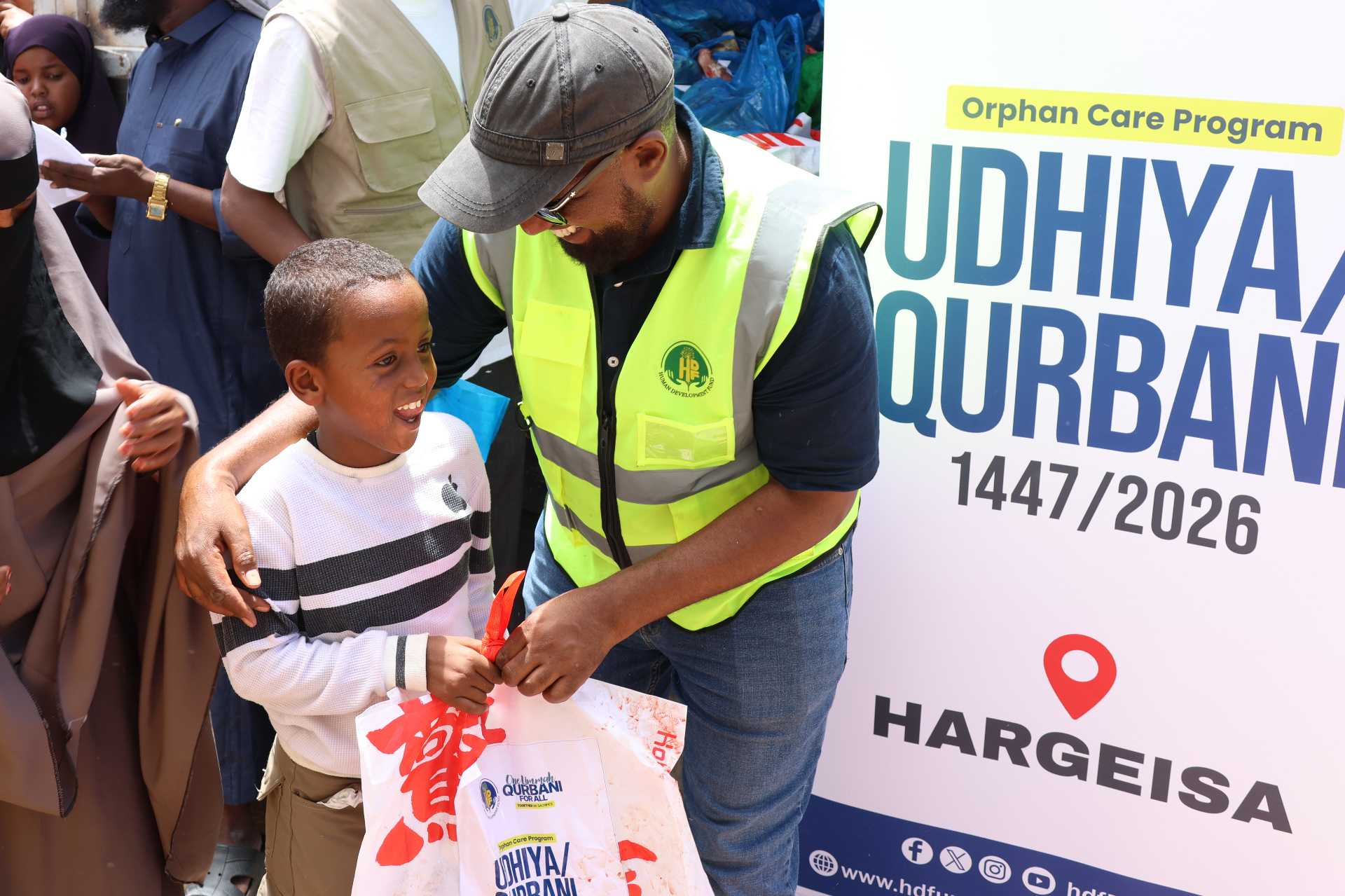 An HDF volunteer in a high-vis vest smiling with a young boy holding a 'One Ummah Qurbani For All' meat share bag in front of the Orphan Care Program Udhiya / Qurbani 1447/2026 Hargeisa banner in Somalia.