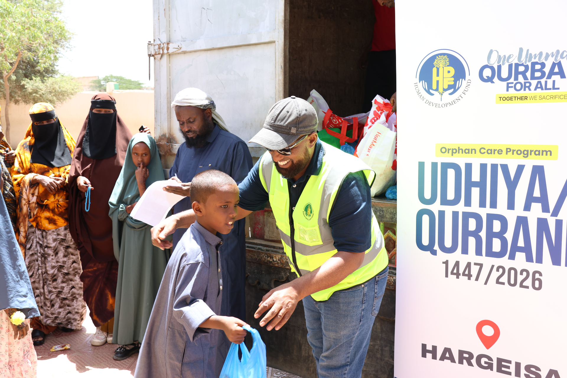 An HDF volunteer in a high-vis vest gently guiding a young boy in a blue thobe holding his Qurbani meat share bag, with families waiting beside the Orphan Care Program Udhiya / Qurbani Hargeisa banner in Somalia.