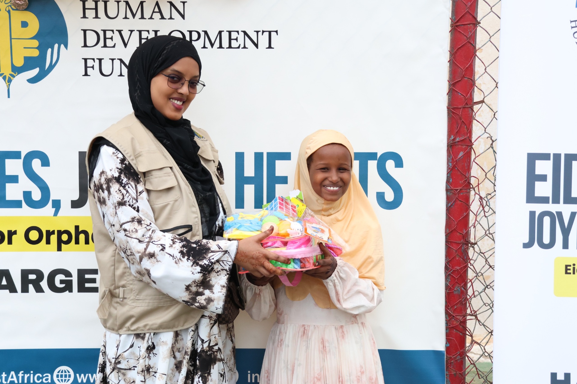 An HDF volunteer handing a basket of Eid gifts to a smiling girl in front of the Eid Smiles, Joyful Hearts banner in Hargeisa, Somalia.