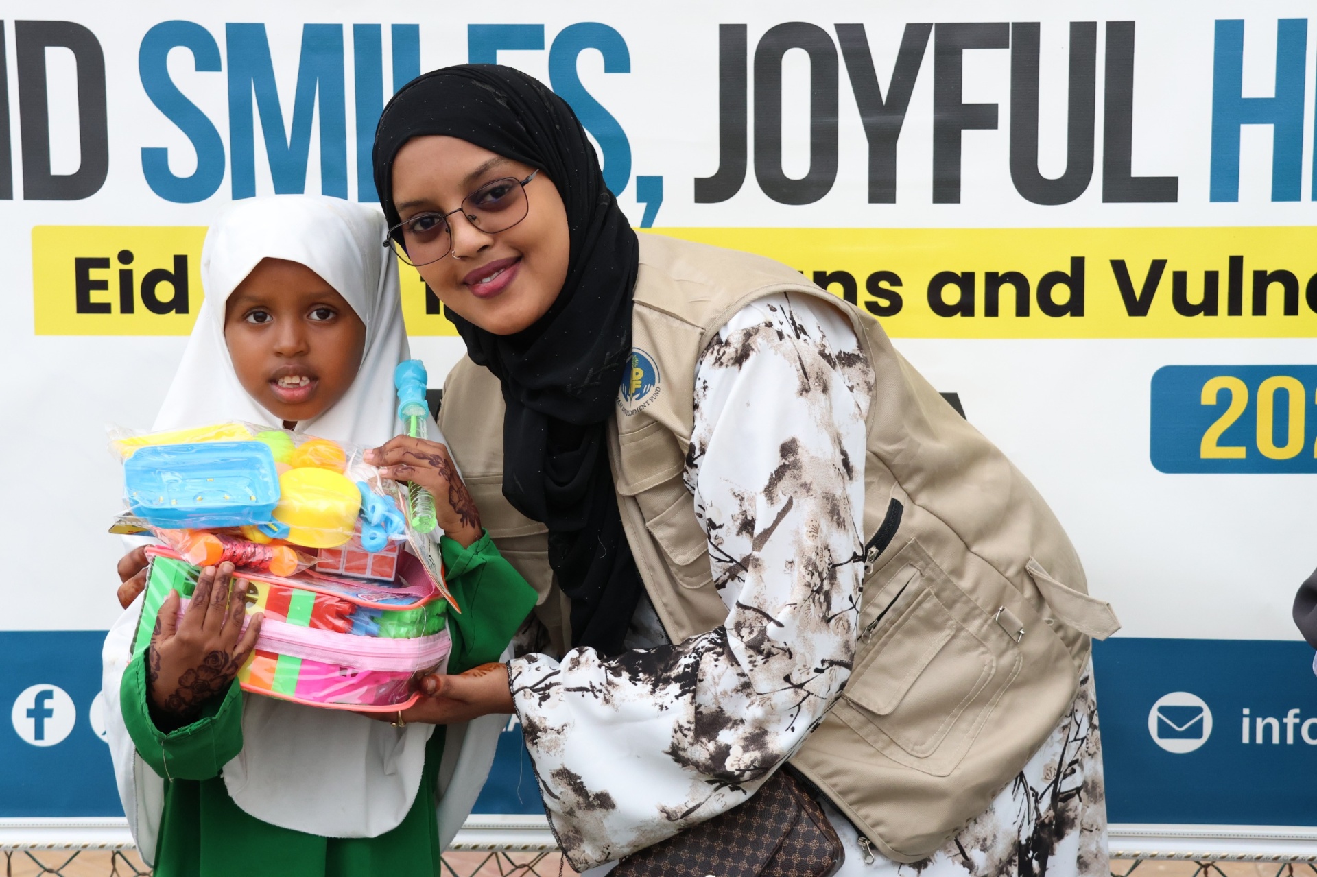 A girl in a green dress holding Eid toys beside a smiling HDF volunteer at the Eid Smiles, Joyful Hearts celebration in Hargeisa, Somalia.