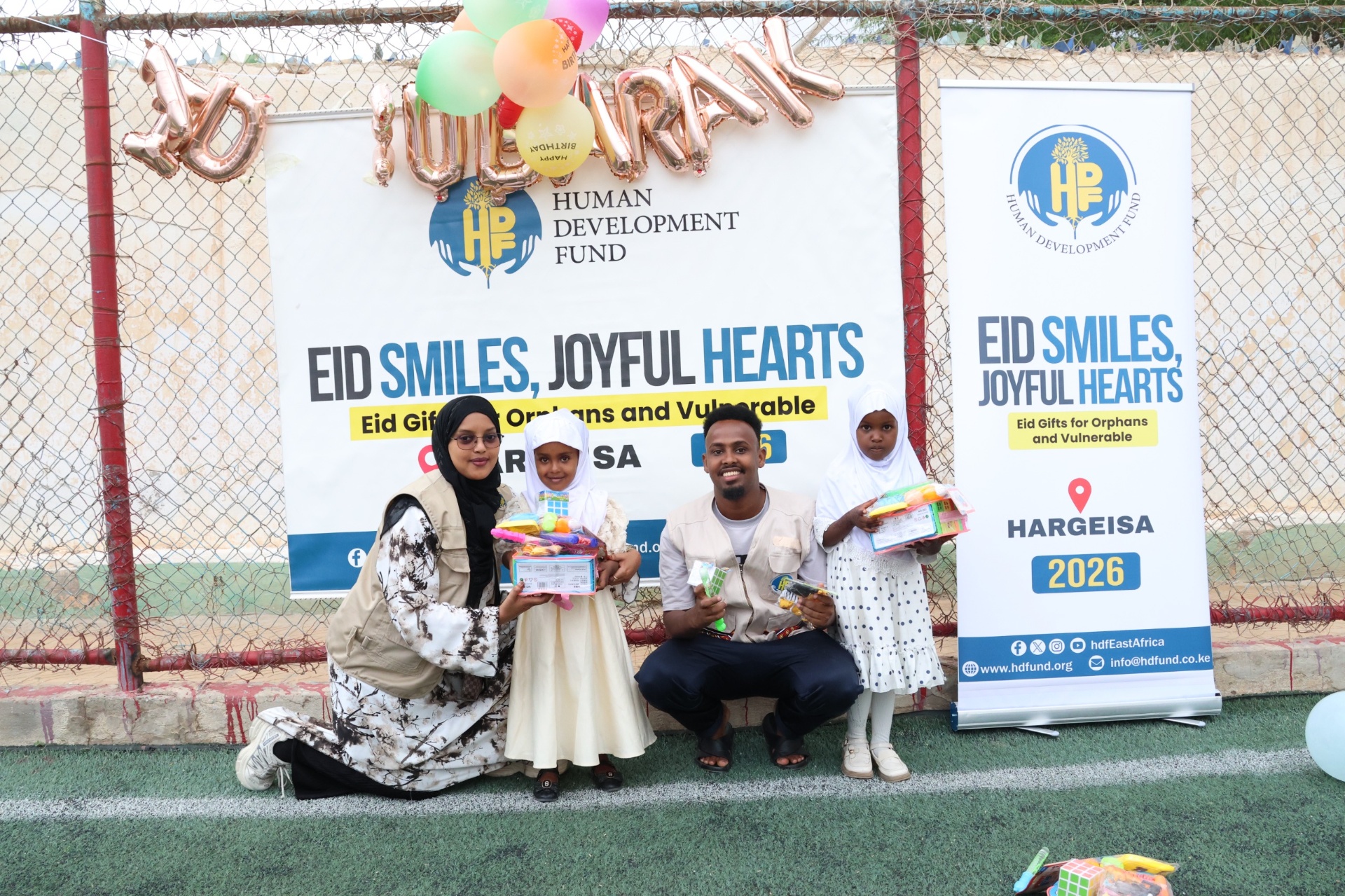 HDF team members posing with two young girls holding Eid gifts in front of the Hargeisa Eid Smiles, Joyful Hearts banner in Somalia.