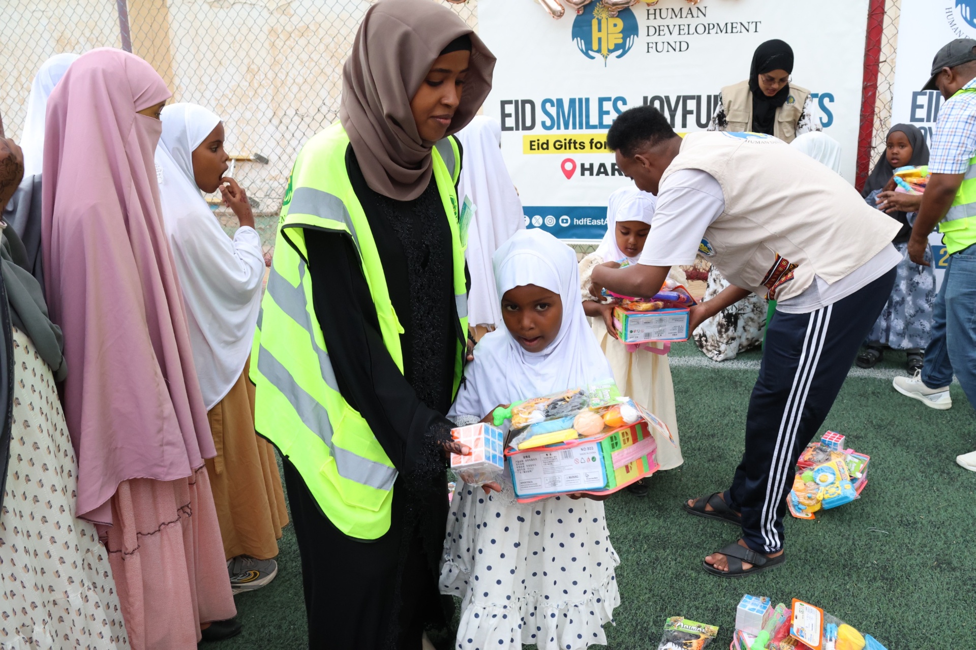 An HDF volunteer in a high-vis vest presenting Eid gifts to a young girl in front of the Hargeisa banner in Somalia.