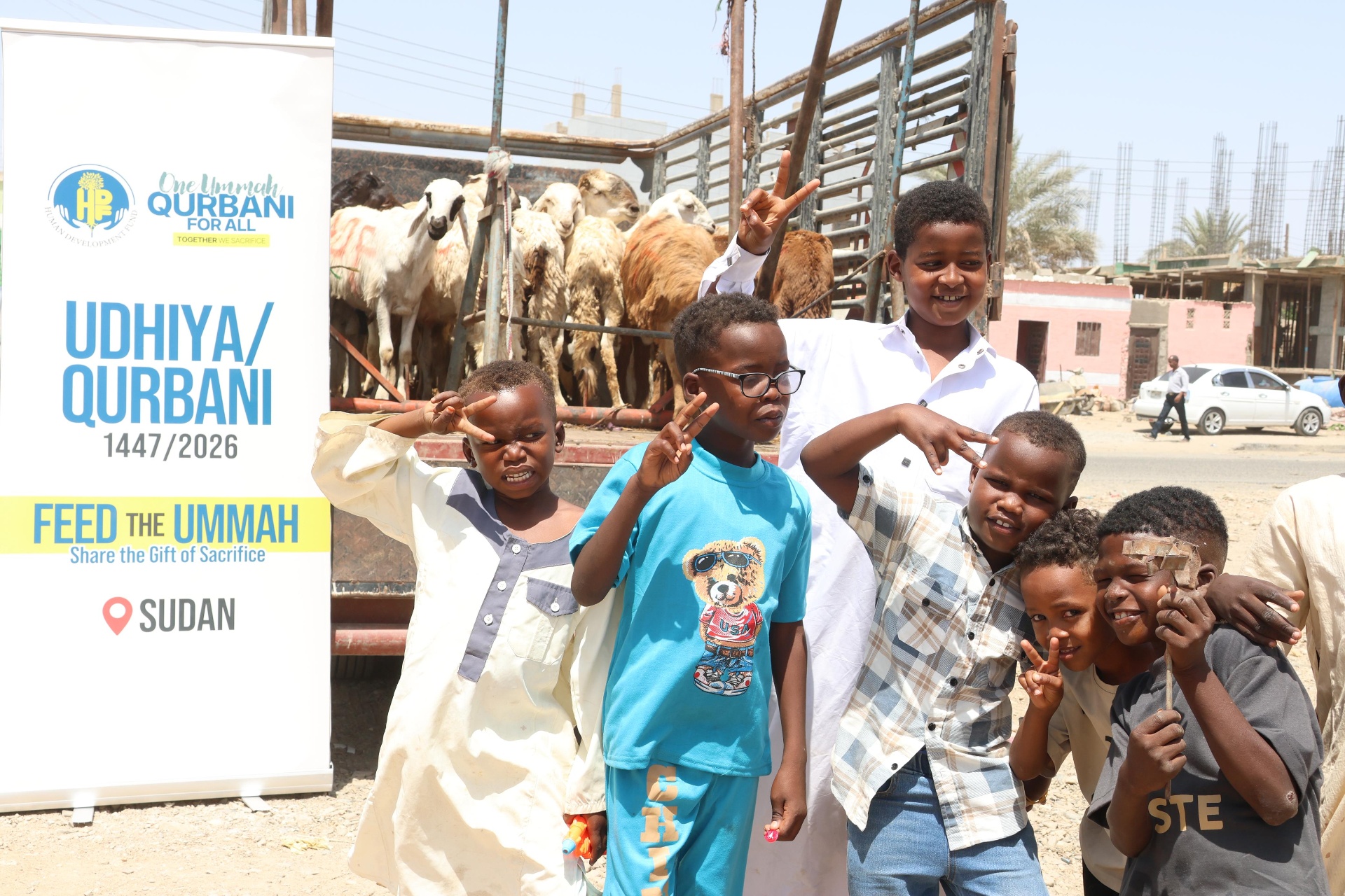 A group of smiling boys throwing peace signs in front of an HDF Qurbani livestock truck and an 'Udhiya / Qurbani — Feed the Ummah, Sudan' banner.