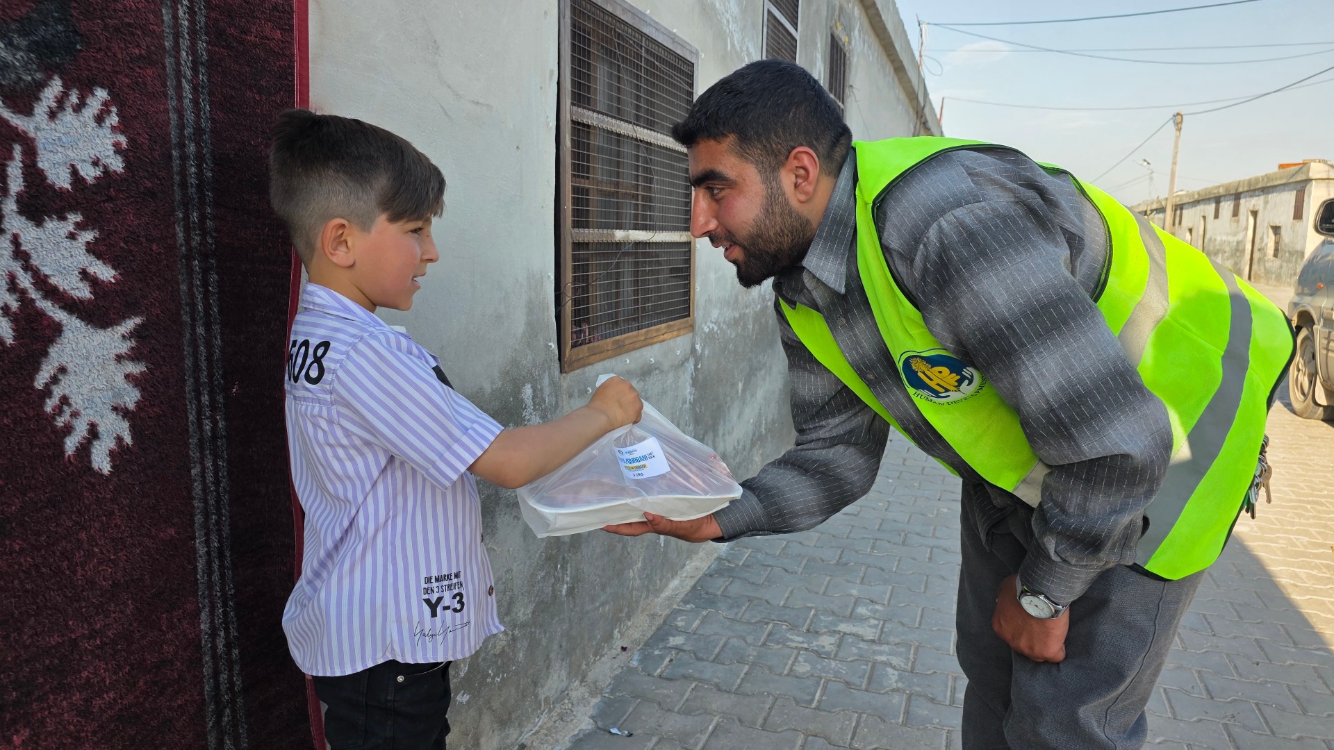 An HDF volunteer in a high-vis vest kneeling to hand a wrapped Udhiya / Qurbani meat share to a smiling young boy at his doorstep in Syria.