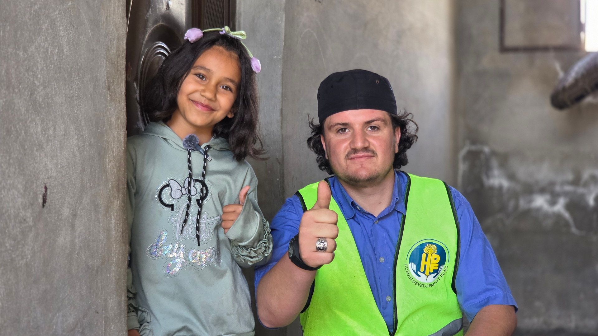 A smiling young girl in a green hoodie standing beside an HDF volunteer in a high-vis vest, both giving a thumbs up after the Qurbani distribution in Syria.