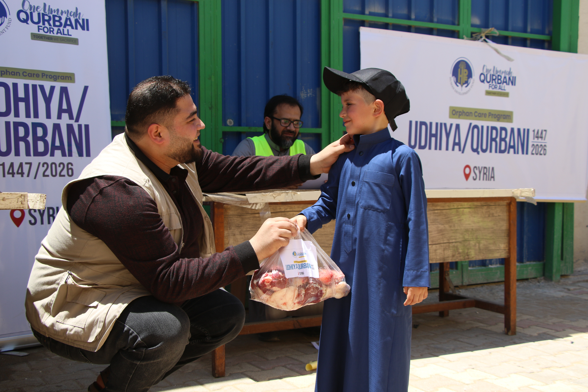 An HDF field worker kneeling to hand a wrapped Udhiya / Qurbani meat share to a young boy in a blue thobe and black cap during the Orphan Care Program distribution in Syria.