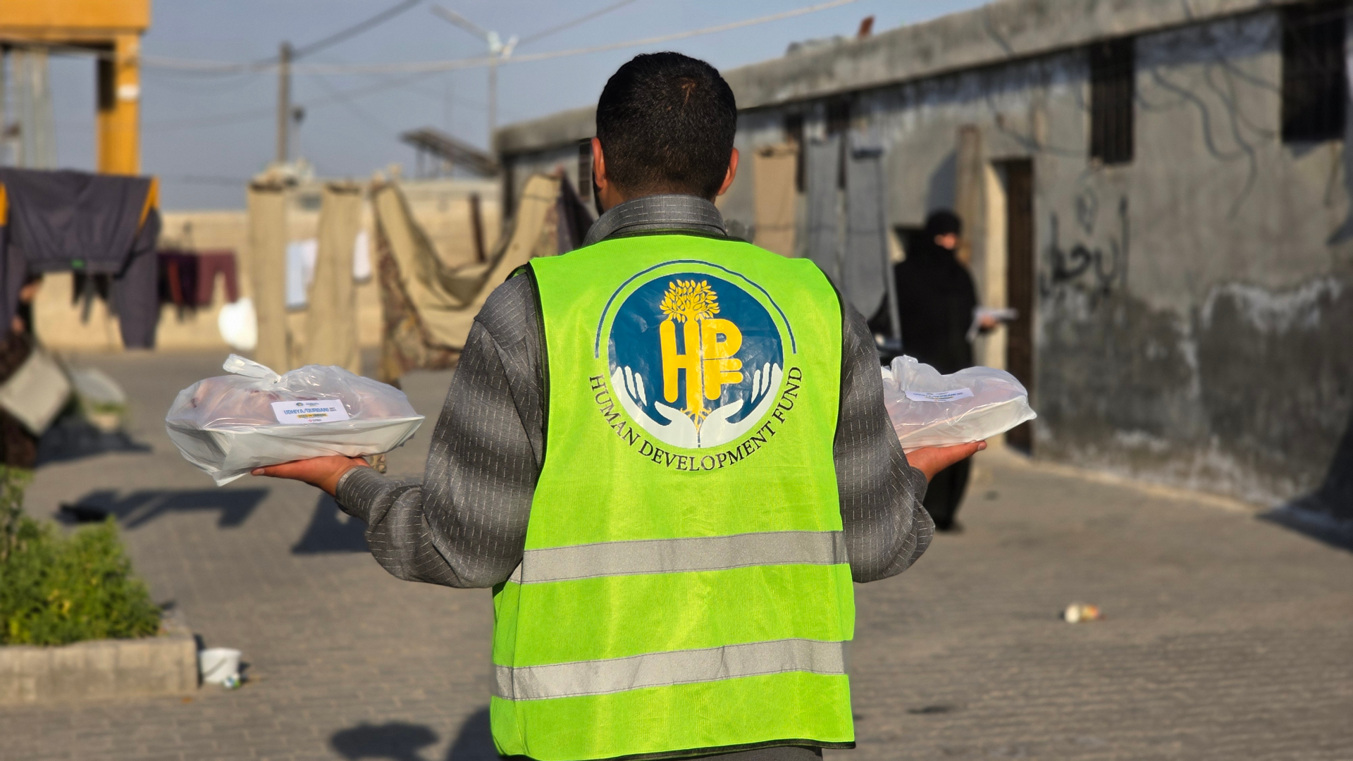An HDF volunteer in a Human Development Fund high-vis vest carrying two wrapped Udhiya / Qurbani meat shares through a displacement camp in Syria.