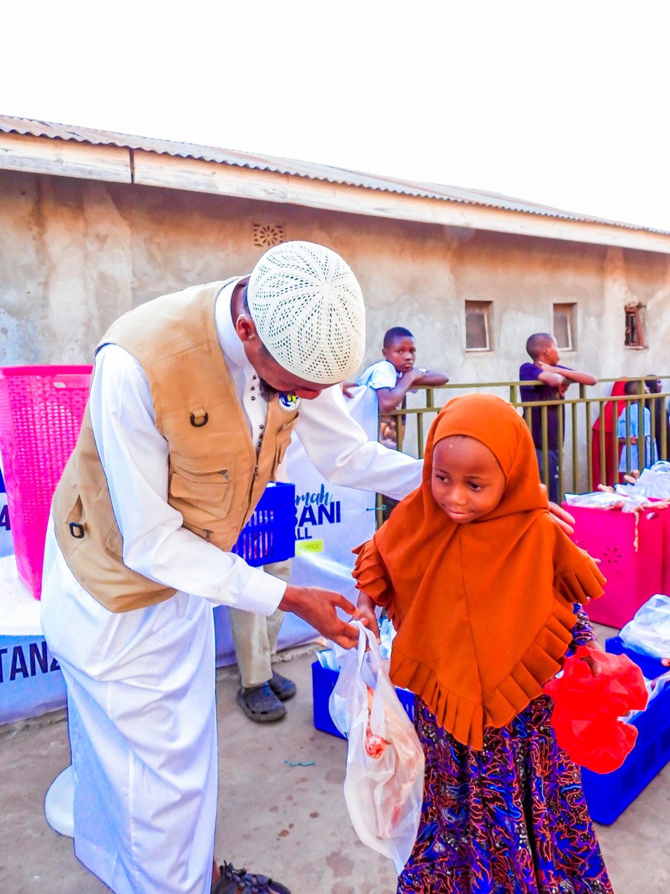 An HDF field coordinator in a white kufi and tan vest gently handing a wrapped Qurbani meat share to a young girl in a bright orange hijab beside a One Ummah Qurbani For All — Tanzania banner.
